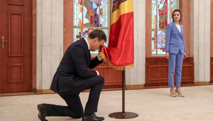 Il neopremier della Moldavia Dorin Recean si inginocchia davanti alla bandiera. Sulla destra, la presidente Maia Sandu